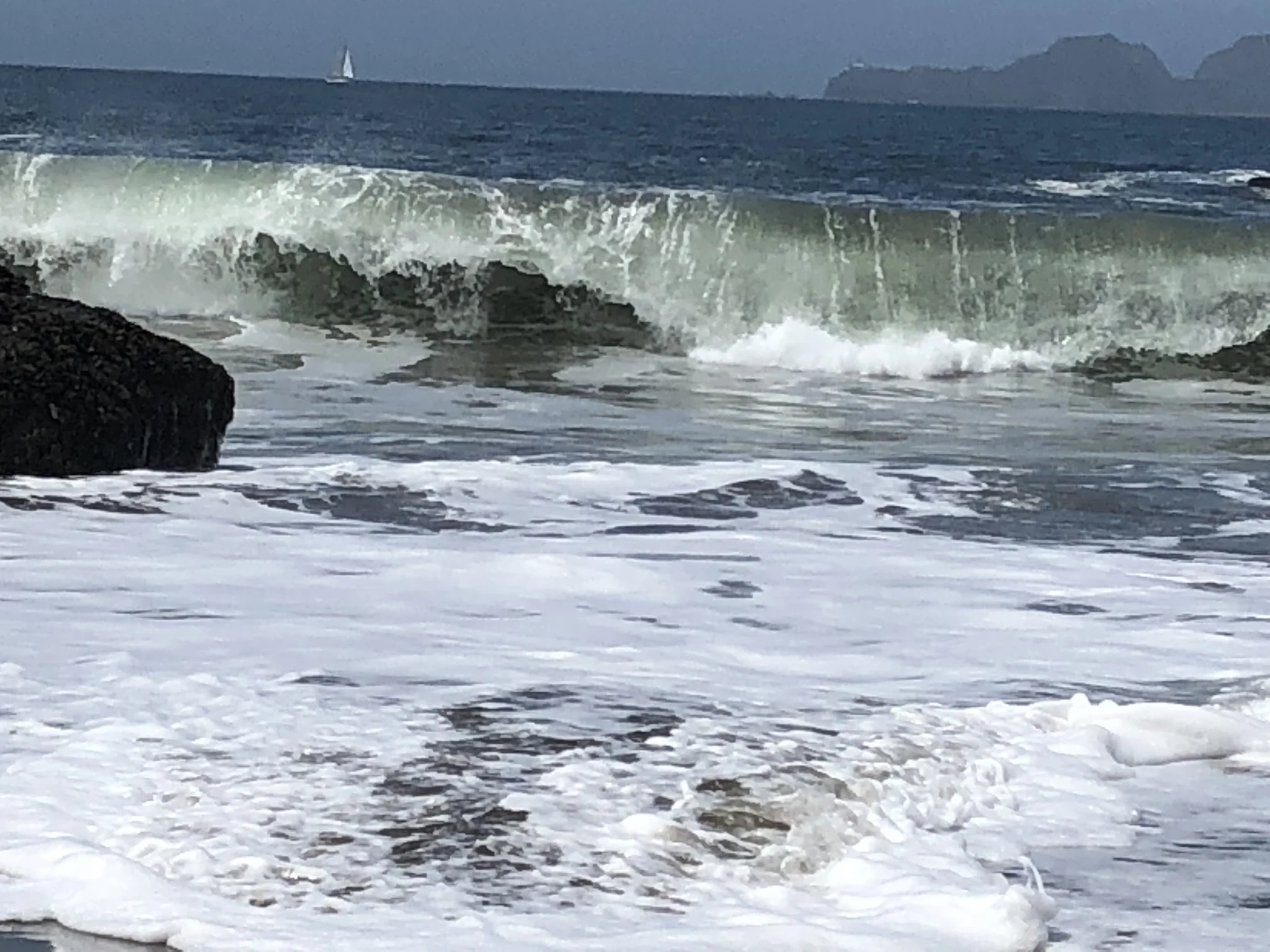 Uma onda se ergue e quebra em espuma sobre as rochas, com o mar aberto ao fundo.