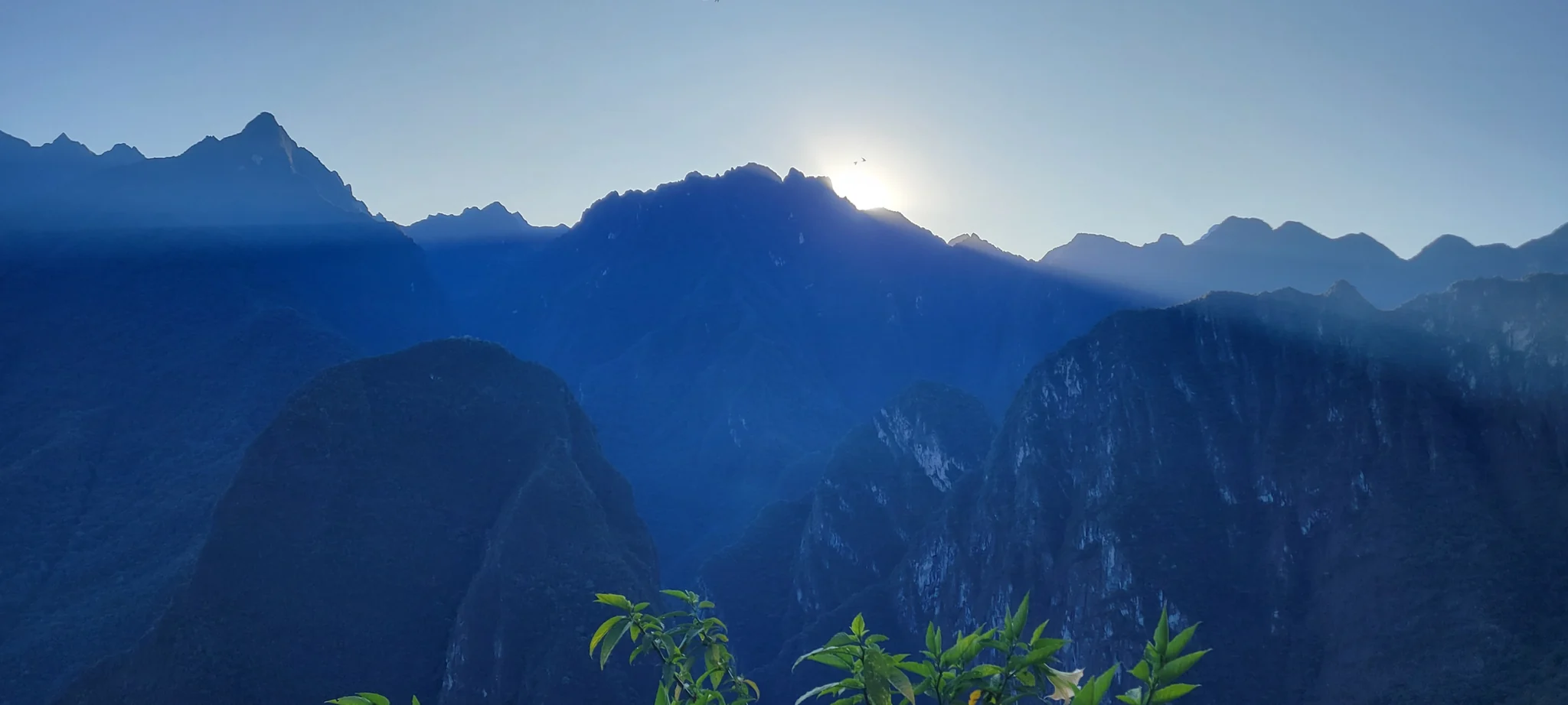 Cordilheira azulada ao amanhecer, com o sol surgindo por trás dos picos.
